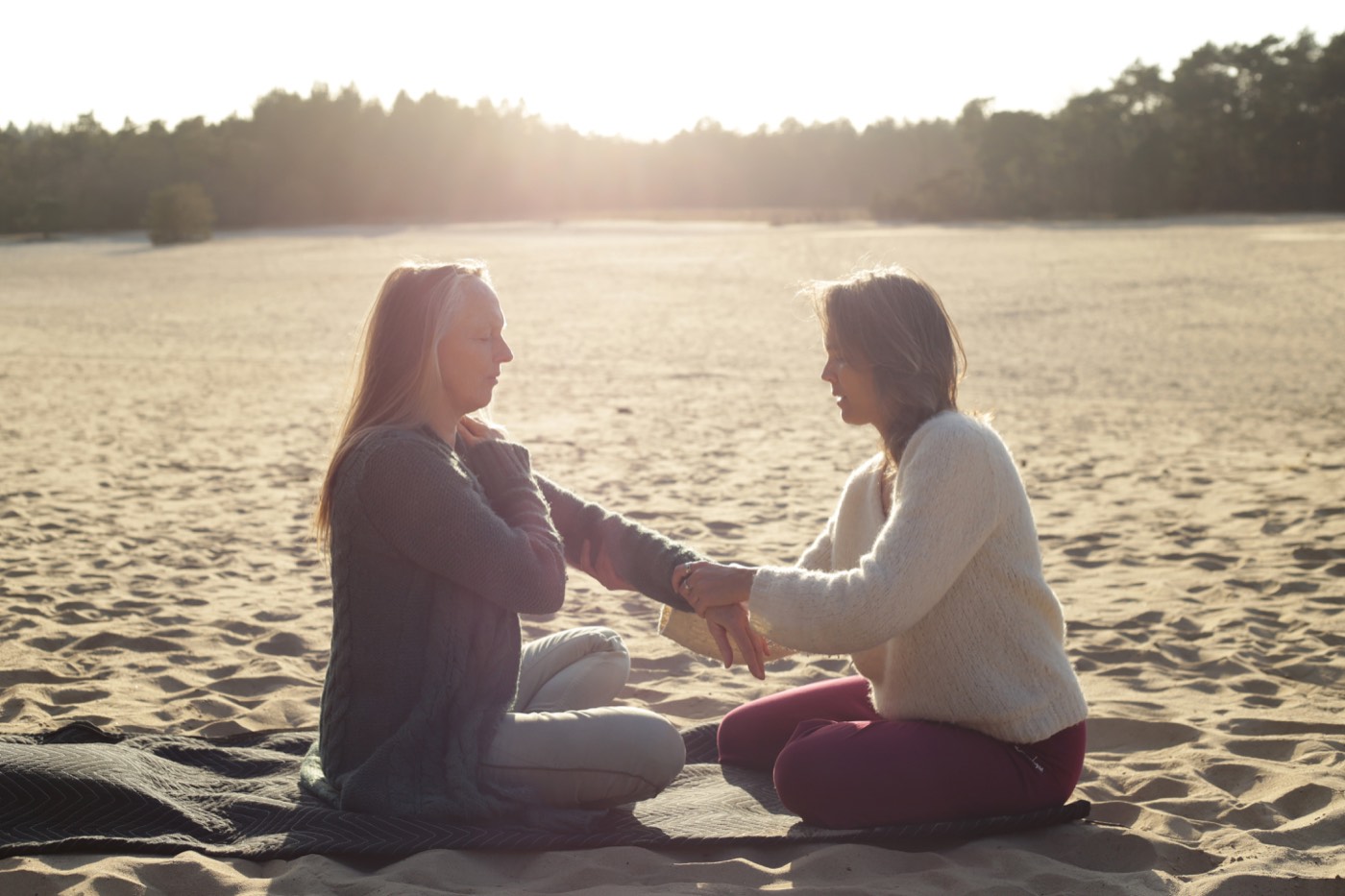 Twee mensen zitten tegenover elkaar in het zand bij zonsondergang, handen die elkaar raken
